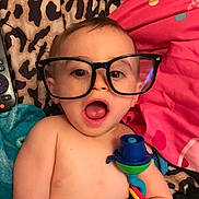 Robbie is registered to the contest to win money with this photo: baby, glasses, toy, blanket, leopard_print, pink_pillow, remote_control, child, shirtless, indoor, cute, surprised, face, head, skin, colorful, playtime, infant, expression, lying_down