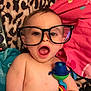 baby, glasses, toy, blanket, leopard_print, pink_pillow, remote_control, child, shirtless, indoor, cute, surprised, face, head, skin, colorful, playtime, infant, expression, lying_down