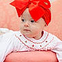 Novella is registered to the contest to win money with this photo: baby, child, red_bow, headband, white_outfit, pink_cushion, blue_eyes, face, portrait, cute, infant, soft_texture, embroidery, curious, indoors, closeup, smiling, sitting, decorative, young