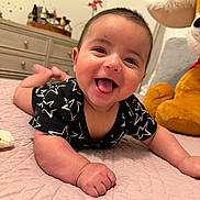 Milan is registered to the contest to win money with this photo: baby, smiling, black_clothing, star_pattern, bedspread, quilted, stuffed_animal, dresser, indoor, child, happy, tongue_out, face, person, cute, infant, furniture, toy, room, lying_down