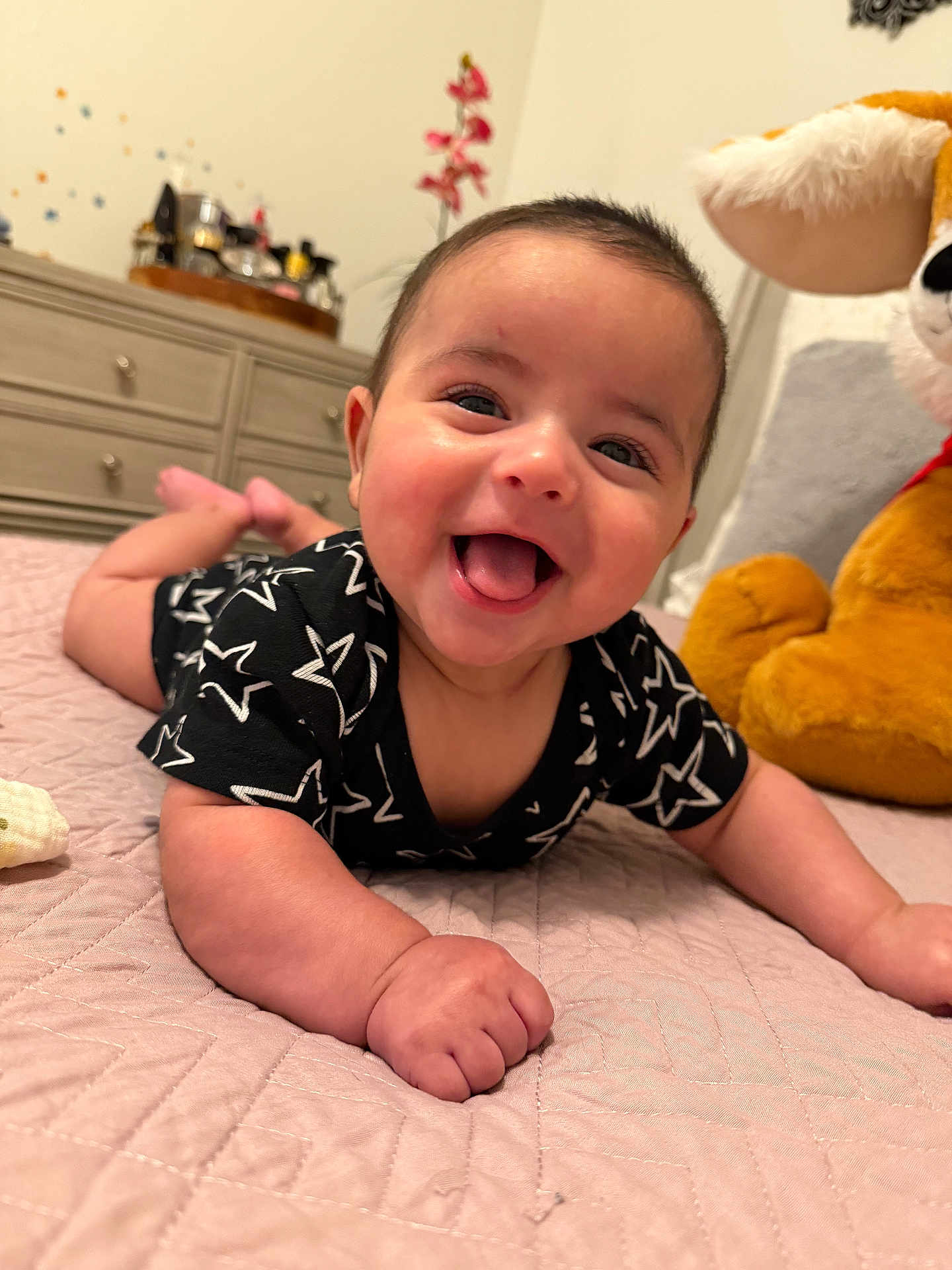 Milan is registered to the contest to win money with this photo: baby, smiling, black_clothing, star_pattern, bedspread, quilted, stuffed_animal, dresser, indoor, child, happy, tongue_out, face, person, cute, infant, furniture, toy, room, lying_down
