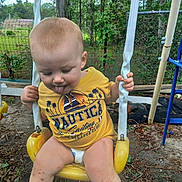 Kayden is registered to the contest to win money with this photo: toddler, child, swing, playground, yellow, outdoor, trees, fence, grass, dirt, clothing, person, tongue_out, play, summer, legs, spots, casual, fun, nature
