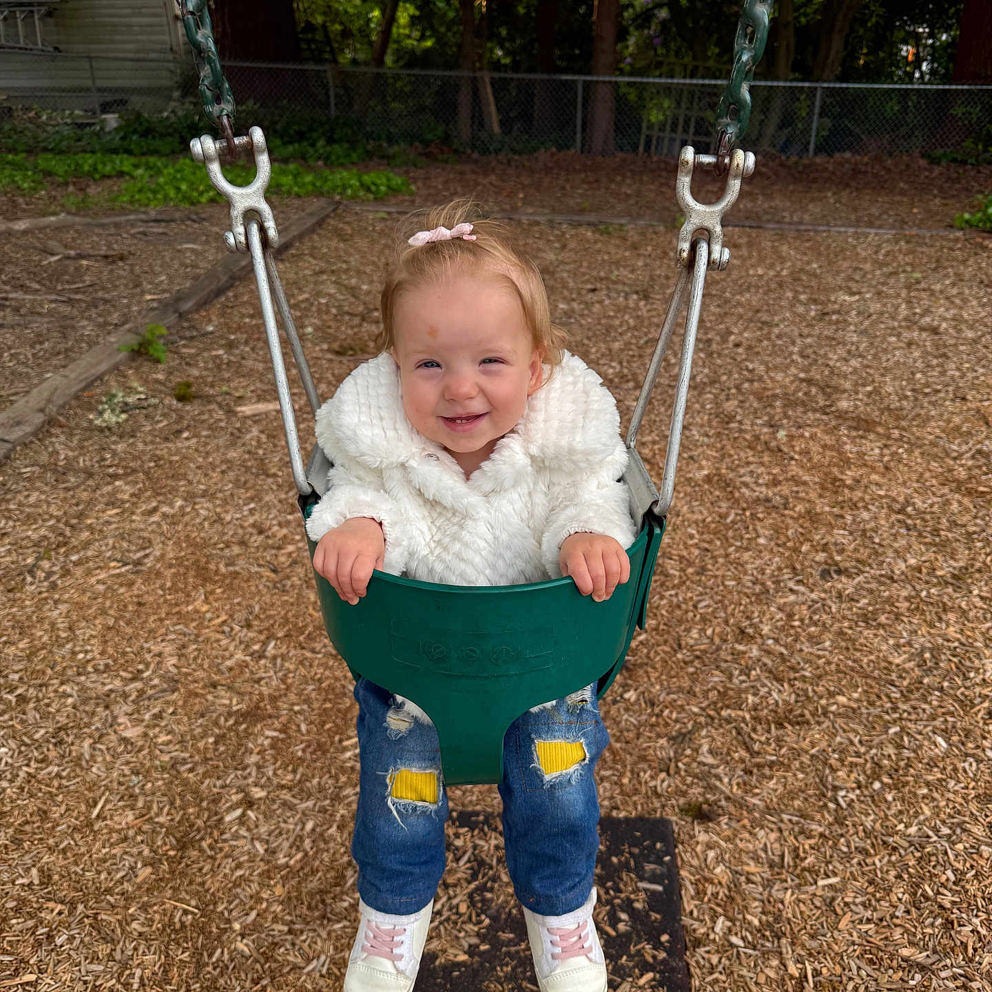 Maggi is registered to the contest to win money with this photo: casual_clothing, child, cute, daylight, fence, fun, happy, jeans, outdoor, park, play_equipment, playground, smiling, sneakers, swing, toddler, trees, white_jacket, wood_chips, young_child