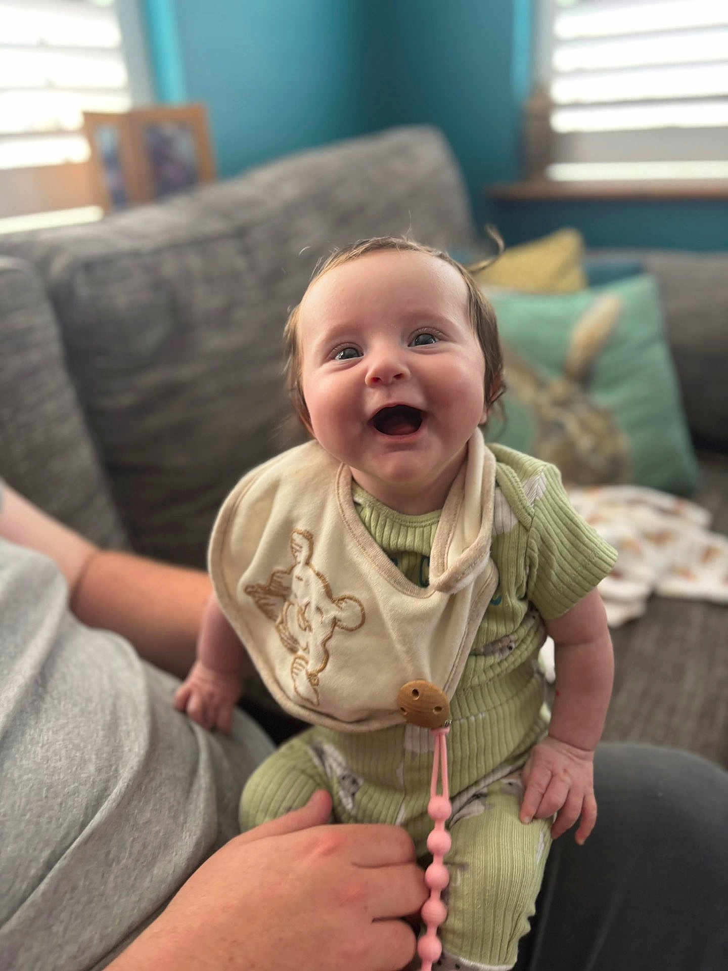 Robyn joined the competition — help win amazing prizes! baby, smiling, infant, person, clothing, bib, hand, couch, cushion, indoor, living_room, window, light, green_outfit, happy, cute, child, seated, adult_hand, home