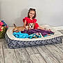 child, dog, pet_bed, blanket, toy, floor, wall, smile, indoor, red_dress, small_dog, cuddle, happy, hair_tied, wooden_floor, cozy, playful, young_girl, domestic, relaxed