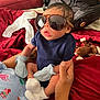 baby, sunglasses, mittens, socks, hand, person, infant, plush_toy, monkey_toy, bed, bedding, red_bedspread, clothing, indoor, portrait, cute, newborn, holding, cozy, casual