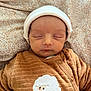 baby, bed, blanket, brown_clothing, child, cozy, cute, face, hat, indoors, infant, newborn, patterned_background, peaceful, portrait, resting, sheep_embroidery, sleeping, soft_texture, warm_colors