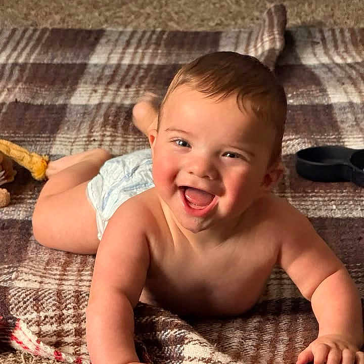 Liam joined the competition — help win amazing prizes! baby, blanket, carpet, child, cute, diaper, expression, face, floor, hand, happy, indoors, infant, lying_down, one_person, plaid, portrait, skin, smiling, young_child