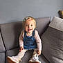 toddler, child, smiling, tongue_out, denim_overall, pink_shirt, tights, sneakers, sitting, couch, gray_couch, hair_clips, indoor, playful, happy, person, furniture, young_child, blonde_hair, casual_clothing