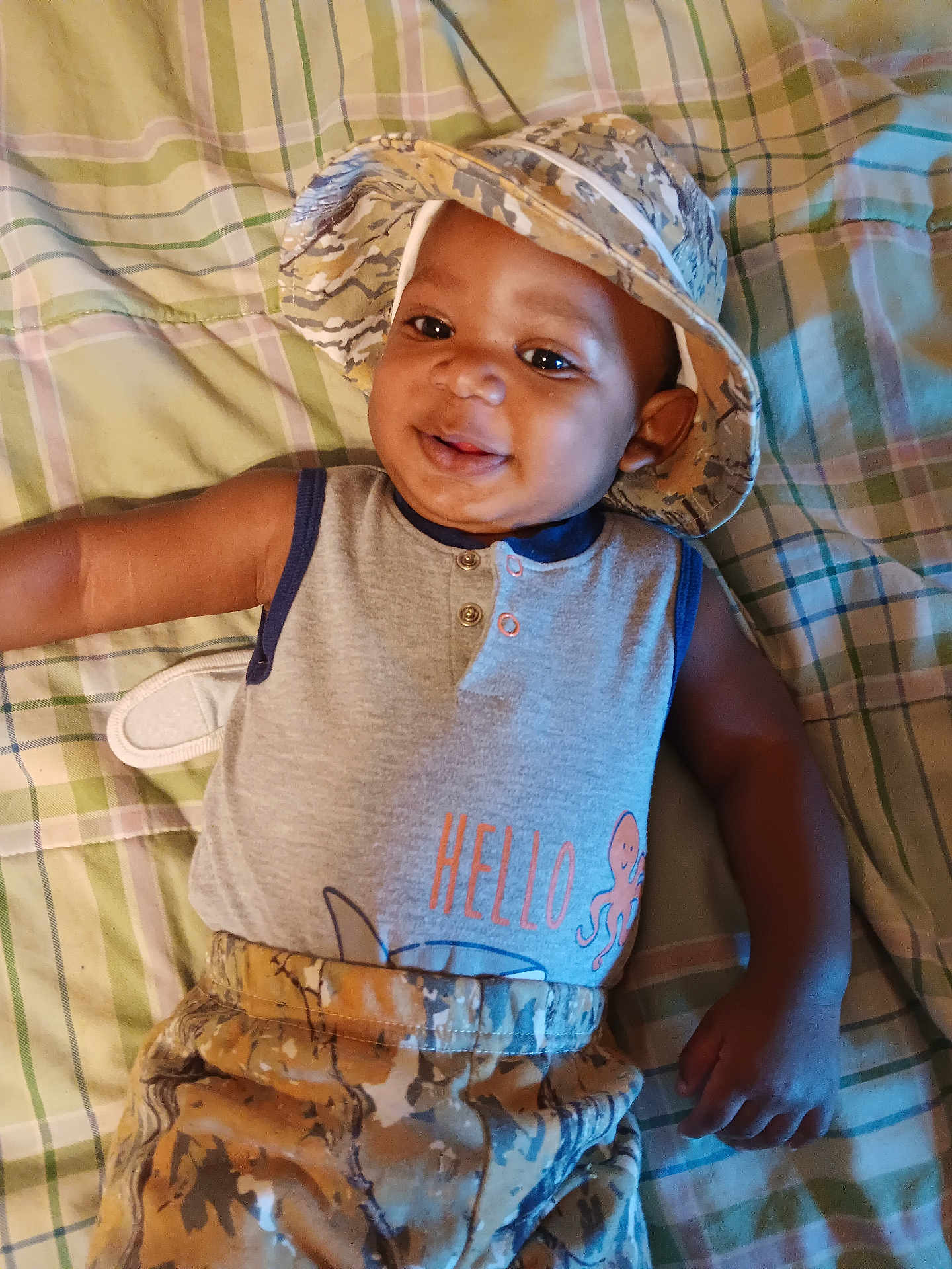 Ja'Juan is registered to the contest to win money with this photo: baby, child, smiling, hat, camouflage, plaid, blanket, sleeveless_shirt, octopus_graphic, casual_clothing, infant, lying_down, indoors, cute, happy, portrait, person, apparel, headwear, patterned_clothing