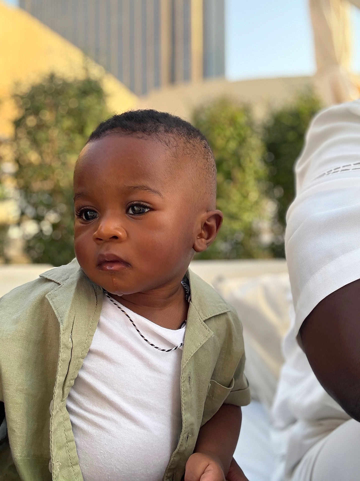 Ahmed a rejoint le concours — aidez-le/la à gagner de superbes lots ! toddler, child, boy, face, portrait, outdoor, urban, casual_clothing, short_hair, necklace, white_shirt, green_jacket, expression, person, young_child, daylight, blurred_background, skin, hand, sitting