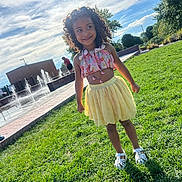 Molli joined the competition — help win amazing prizes! child, girl, curly_hair, smile, yellow_skirt, floral_top, grass, outdoor, sunny, water_fountain, park, playful, happy, summer, nature, casual_clothing, shoes, greenery, blue_sky, clouds