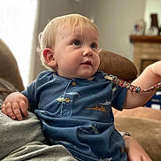 Levi joined the competition — help win amazing prizes! toddler, child, blue_shirt, dinosaur_pattern, indoor, living_room, curious, seated, person, adult_hand, couch, soft_lighting, blond_hair, young_child, casual_clothing, portrait, home_interior, relaxed, cute, family_moment