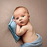 baby, infant, blue_pillow, blanket, resting, curious_expression, wide_eyes, soft_texture, beige_background, skin, newborn, portrait, lying_down, cute, child, peaceful, tender, wrapped, face, head
