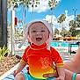 baby, child, smiling, hat, poolside, swimwear, sunhat, outdoor, summer, palm_trees, blue_sky, water, towel, person, happy, vacation, sunny, sitting, bright_colors, daytime