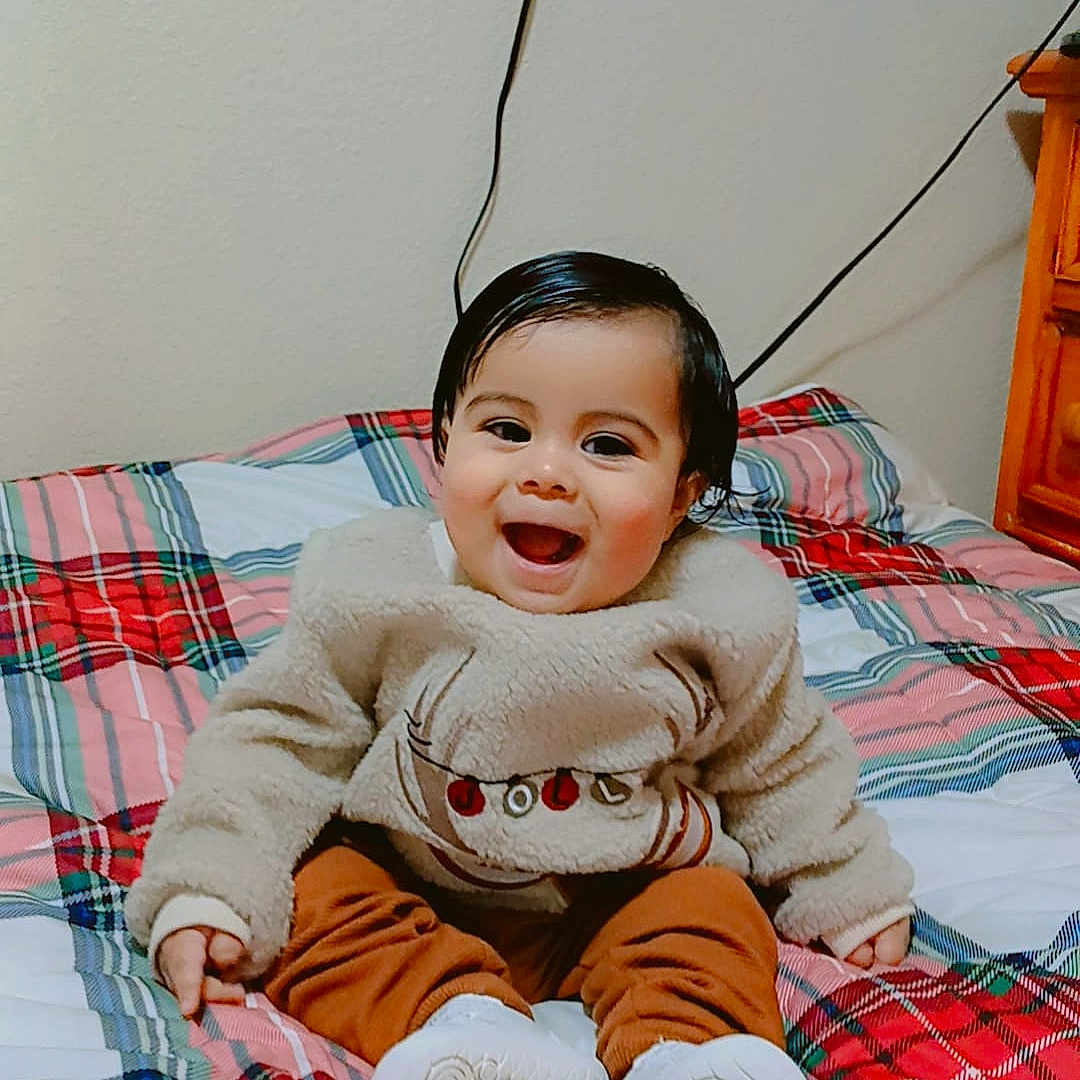 Roberth is registered to the contest to win money with this photo: baby, bed, blanket, cable, child, cozy, cute, furniture, happy, indoor, pants, person, plaid, portrait, shoes, sitting, smiling, sweater, television, wall