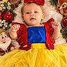baby, child, snow_white_costume, portrait, lying_down, smiling, face, red_bow, blue_dress, yellow_skirt, fairy_tale, roses, apple, stuffed_animal, bird, dwarf_toy, lantern, blanket, floral_decoration, indoor