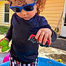 child, toddler, curly_hair, sunglasses, toy_car, rubber_duck, swim_trunks, kiddie_pool, water, summer, outdoor, play, yellow_house, sunny_day, striped_clothing, blue_shirt, hands, concrete, door, window
