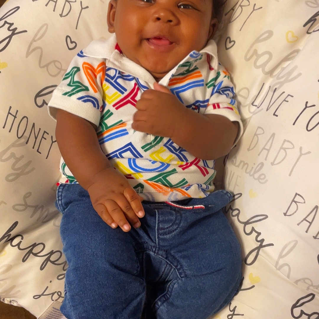 Jakai is registered to the contest to win money with this photo: baby, infant, smiling, child, clothing, jeans, colorful_shirt, pillow, cushion, text, happy, cute, indoors, person, skin, face, foot, hand, sitting, portrait