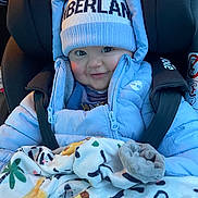 Elio participe au concours pour gagner de l'argent avec cette photo : baby, child, car_seat, winter_clothing, puffer_jacket, beanie, blanket, animal_print, smile, face, cute, blue, warm, infant, cozy, sitting, person, clothing, headwear, indoors