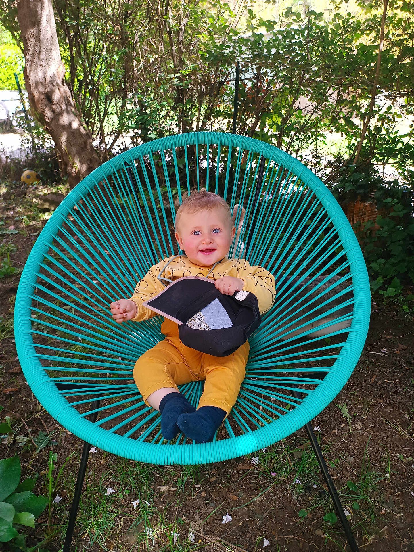 Noé a rejoint le concours — aidez-le/la à gagner de superbes lots ! baby, child, smiling, blue_eyes, yellow_clothing, turquoise_chair, outdoor, greenery, grass, tree, cap, sitting, happy, nature, sunlight, portrait, young_child, cute, playful, garden
