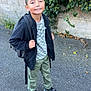 child, boy, smiling, backpack, jacket, pants, sneakers, ivy, stone_wall, outdoor, sidewalk, greenery, nature, casual_clothing, portrait, standing, happy, person, daylight, urban