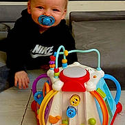 Lylio a rejoint le concours — aidez-le/la à gagner de superbes lots ! activity_center, baby, bright_colors, child, colorful_toy, cushion, face, hand, hoodie, indoor, nike_logo, pacifier, playtime, portrait, sitting, smiling, sofa, toddler, toy, wooden_floor