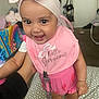 baby, child, pink_bib, headband, smiling, indoor, person, happy, clothing, footwear, hand, blanket, baby_clothes, toy, carpet, room, face, cute, infant, holding