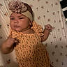 Arianna is registered to the contest to win money with this photo: baby, infant, child, portrait, lying_down, crib, mattress, floral_pattern, headband, flower, dress, orange_clothing, frills, face, eyes, arms, legs, socks, indoors, sleeping_area