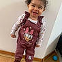 toddler, child, smiling, curly_hair, indoor, overalls, daisy_duck, boots, wooden_floor, wall, standing, happy, clothing, person, footwear, cute, young_child, playful, portrait, fashion
