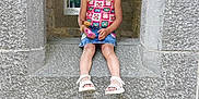 Lilya participe au concours pour gagner de l'argent avec cette photo : child, girl, stone_wall, archway, window, toy, playful_expression, colorful_clothing, denim_skirt, sandals, outdoor, nature, greenery, historic_architecture, sitting, portrait, young_child, casual, smile, stone_texture