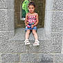 child, girl, stone_wall, archway, window, toy, playful_expression, colorful_clothing, denim_skirt, sandals, outdoor, nature, greenery, historic_architecture, sitting, portrait, young_child, casual, smile, stone_texture