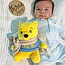 baby, infant, plush_toy, yellow_bear, knitted_blanket, wooden_sign, three_months, onesie, striped_clothing, scarf, soft_toy, child, portrait, laying_down, cute, indoor, calm, curious, baby_clothes, baby_boy