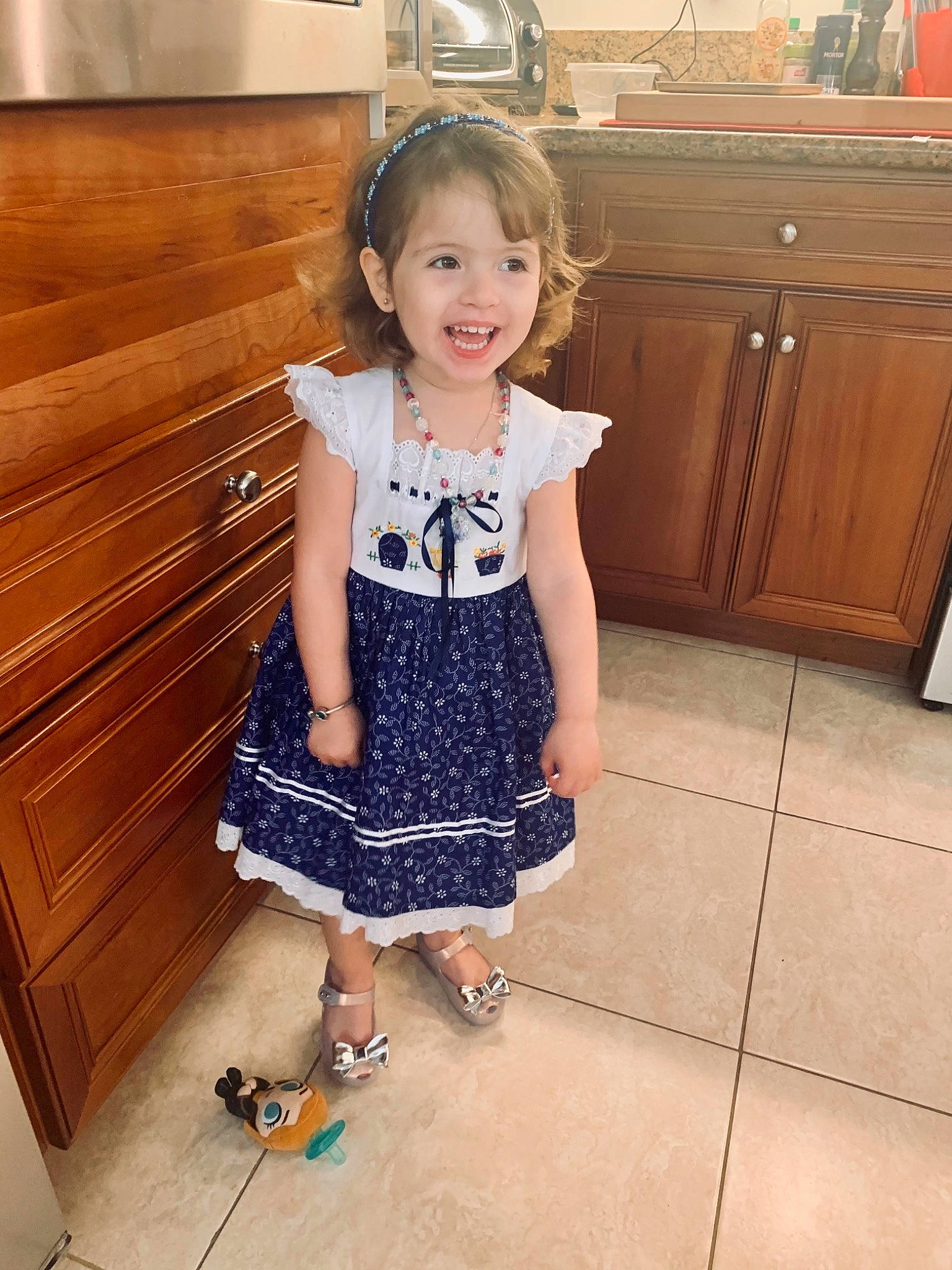 Beatriz is registered to the contest to win money with this photo: baby_toddler_clothing, cabinetry, countertop, drawer, dress, floor, flooring, fun, happy, home_appliance, joy, kitchen_appliance, person, sleeve, smile, stairs, standing, thigh, toddler, trunk