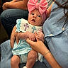 baby, infant, pink_bow, headband, blue_outfit, cross_embroidered, holding_hands, adult, jeans, seated, curious_expression, indoor, casual_clothing, white_shoes, close_up, portrait, person, family, cute, hands