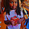 child, girl, portrait, smiling, braids, curly_hair, pizza, food, eating, indoors, party, birthday, t_shirt, shorts, standing, wooden_floor, chairs, background_people, colorful_lighting, casual_clothing