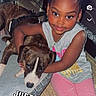 child, girl, dog, pet, hugging, smiling, portrait, indoors, sitting, casual_clothing, pink_pants, gray_shirt, necklace, curly_hair, brown_dog, white_markings, couch, home, social_media, text_overlay