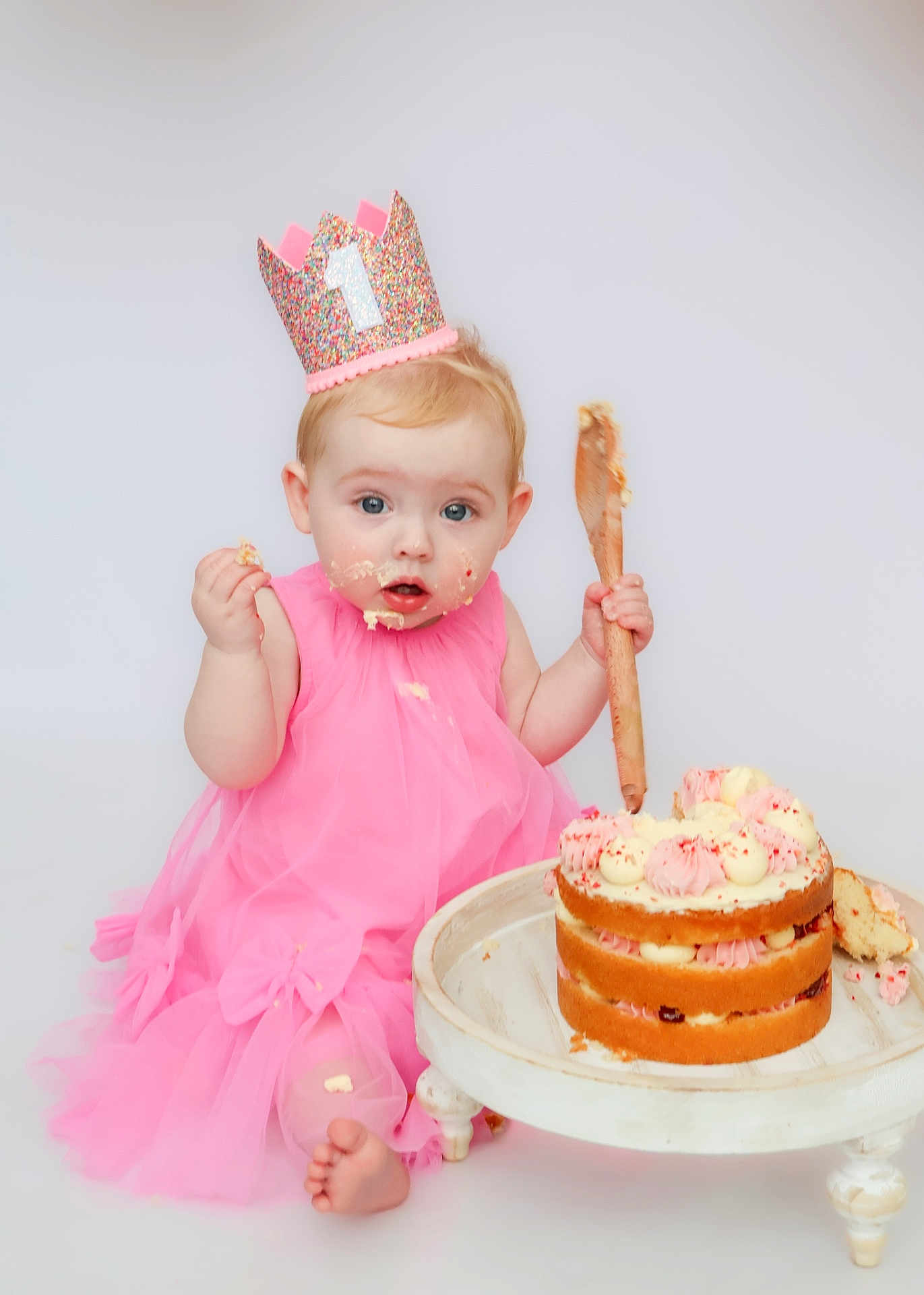 Grace is registered to the contest to win money with this photo: baby, birthday, cake, pink_dress, crown, frosting, wooden_spoon, celebration, party, child, smudged_face, sitting, foot, layered_cake, white_background, cute, messy, food, happy, indoor