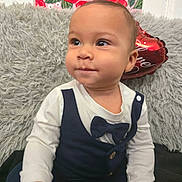 Liam is registered to the contest to win money with this photo: baby, infant, toddler, bow_tie, onesie, heart_balloon, decorative_balloon, floral_wallart, fuzzy_blanket, pillow, portrait, sitting, cute, cheeks, eyes, hands, indoor, clothing, smile, photography