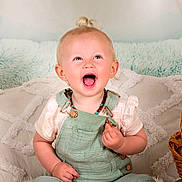 Kimber is registered to the contest to win money with this photo: child, toddler, baby, smiling, laughing, open_mouth, blond_hair, top_knot, earrings, necklace, green_overalls, striped_shirt, wicker_basket, blanket, decorative_pillow, indoor, portrait, sitting, joyful, soft_lighting