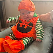 Tommy a rejoint le concours — aidez-le/la à gagner de superbes lots ! baby, child, pumpkin_costume, orange, green_stripes, striped_clothing, cute, infant, costume, smiling, sitting, hand, adult, cushion, indoors, soft_light, portrait, face, expression, holiday