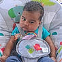 baby, child, infant, curly_hair, pacifier, baby_seat, colorful, animal_print, toy, clothing, blue_shirt, pants, straps, indoors, seat, cute, expression, face, person, sitting