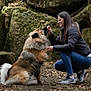 woman, dog, outdoor, forest, rocks, leaves, crouching, feeding, pet, fur, casual_clothing, glasses, long_hair, sneakers, nature, friendship, animal, brown, black, white