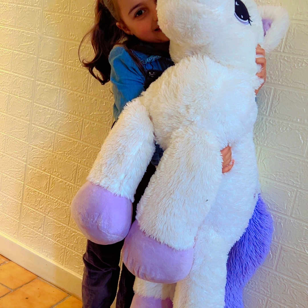 Kenza a rejoint le concours — aidez-le/la à gagner de superbes lots ! casual_clothing, child, childhood, cute, girl, hair_accessory, happy, home, indoor, pink_shoes, playful, plush_toy, purple, smiling, soft_toy, stuffed_animal, tile_floor, toy, unicorn, wall