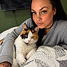 woman, cat, calico_cat, bed, quilt, indoor, portrait, snuggling, cozy, casual_clothing, long_hair, green_eyes, face, pet, animal, resting, blanket, choker_necklace, relaxed, human