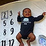 baby, black_onesie, blanket, celebration, child, cute, face, feet, happy, head, indoor, infant, legs, lying_down, milestone, numbers, person, raised_arms, skin, smiling
