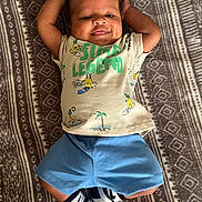 Adonis is registered to the contest to win money with this photo: baby, child, smiling, lying_down, tshirt, shorts, socks, blanket, pattern, happy, cute, indoors, person, face, head, arms, legs, footwear, infant, casual_clothing