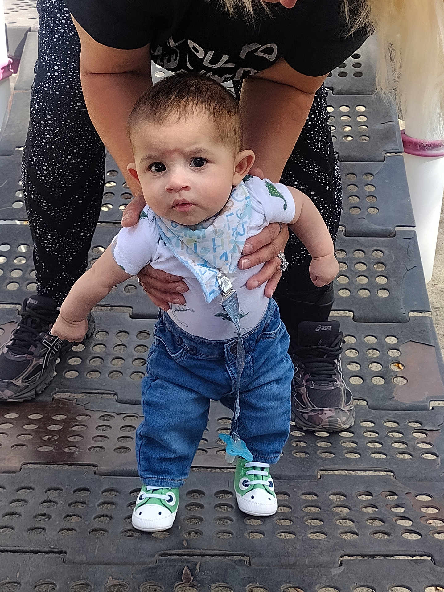 Adam is registered to the contest to win money with this photo: baby, child, adult, hands, jeans, bib, shoes, sneakers, outdoor, playground, standing, support, person, clothing, expression, face, footwear, textured_surface, casual_wear, dinosaur_shoes