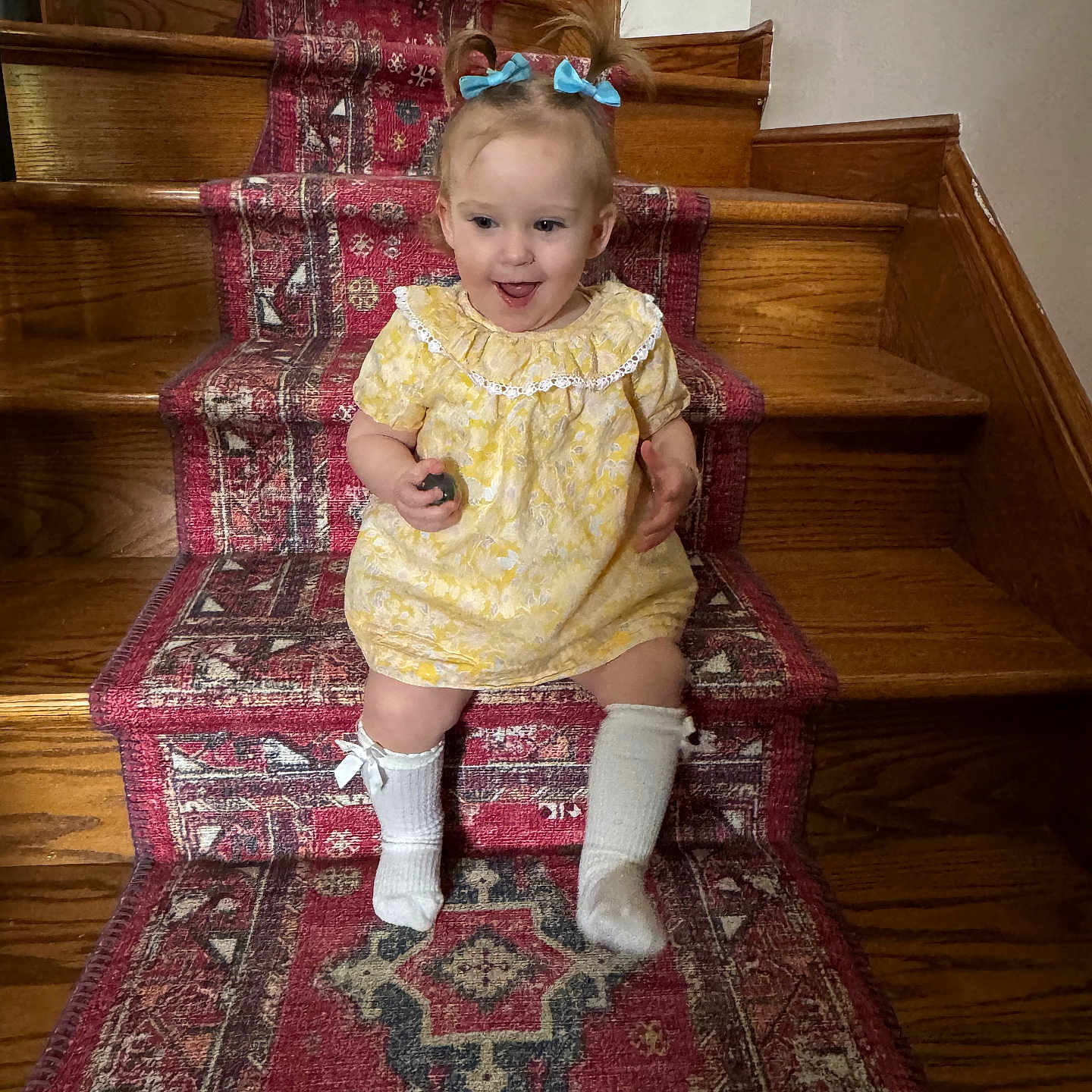 Theadora is registered to the contest to win money with this photo: baby, blue_bows, carpet, child, cute, hair_tied, happy, holding_object, home, indoor, patterned_rug, person, playful, sitting, smiling, stairs, toddler, white_socks, wooden_stairs, yellow_dress