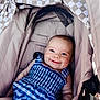 baby, smiling, stroller, blue_clothing, checkered_pattern, infant, child, happy, cute, drool, outdoor, seat, fabric, canopy, person, young_child, portrait, resting, casual, comfort
